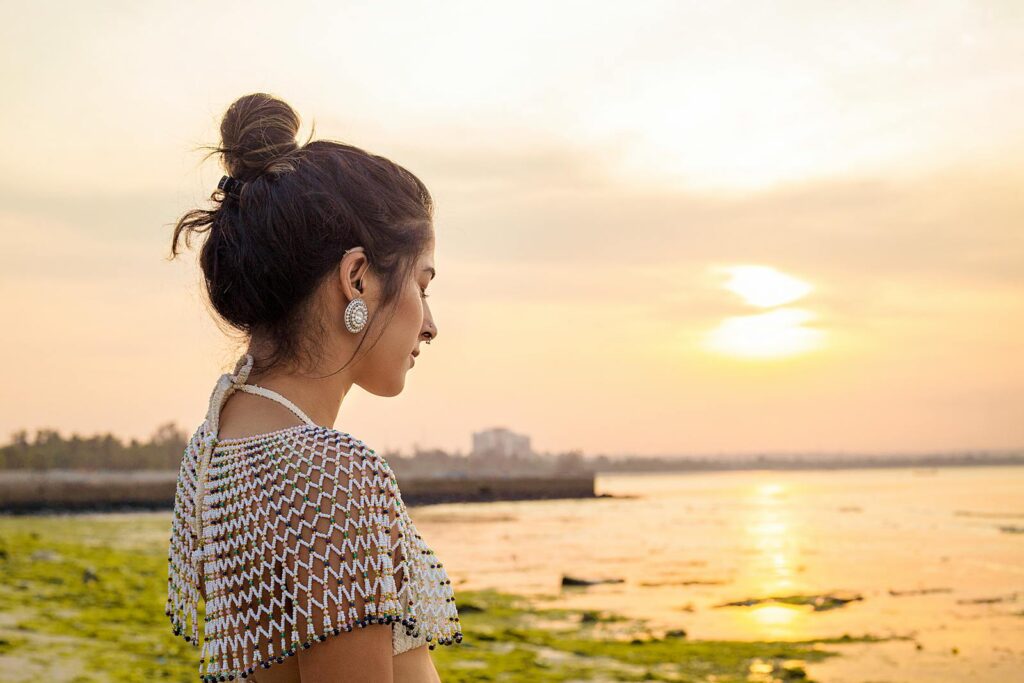 Calm ethnic female with brown hair standing with closed eyes on seashore with green algae in sunny day